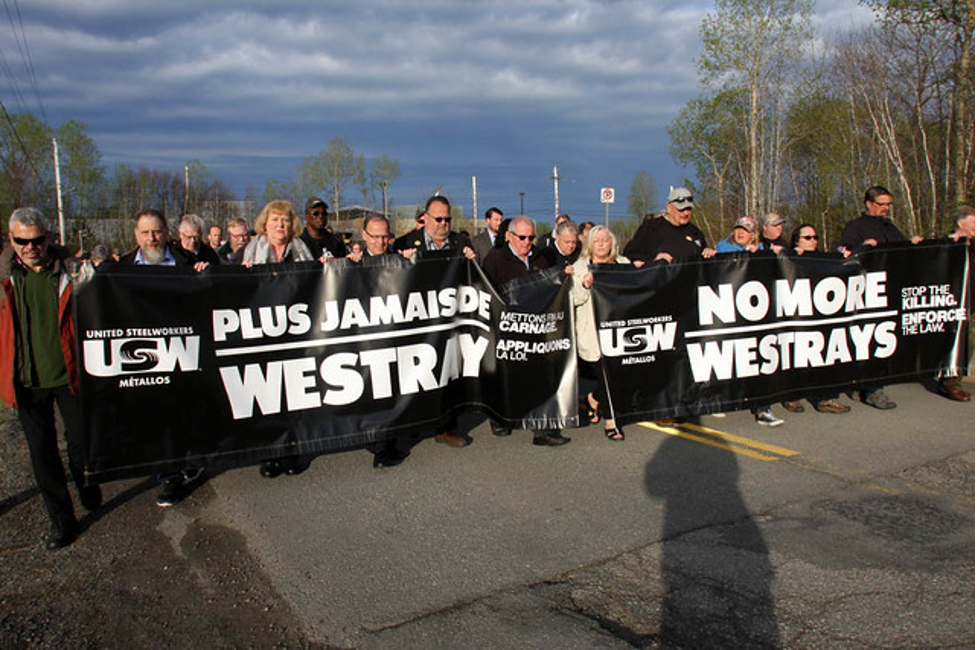Image: A group of Steelworkers march down a street carrying two black banners with white lettering - Plus jamais de Westray / No more Westrays