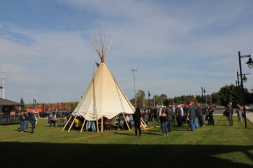 Image: A group of people standing outside. Everyone is surround a traditional teepee.