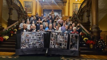 Rows of people standing on the large staircase in the legislature. There are 7 people at the very front holding a banner with photos of various miners who were affected by McIntyre Powder. Text at the bottom of the banner says 