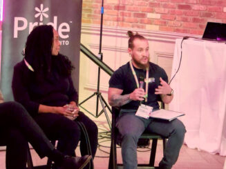 Image: Two people are sitting on chairs as part of a group discussion. A bearded white man in a USW t-shirt with tattoos on his forearn is holding a microphone and speking, gesturing with his hand as he talks. He has a notebook on his lap. A Black woman with long dark hair to his right is observing as he speaks. She is holding a glass of water. A Pride Toronto standup banner is behind them. They are in a room with red brick walls. A computer is on a high table off to the side. There is other meeting equipment behind the speakers - a light stand and a long black cord from the computer.