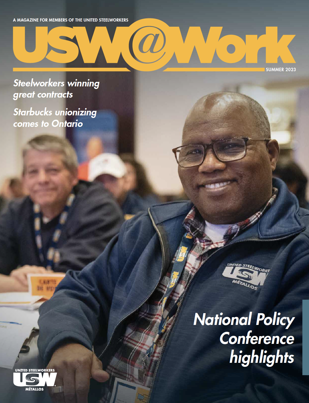 Image: cover of a magazine featuring a smiling black man who is seated at a union event in the foreground. He is wearing a USW jacket and smiling. Behind him in the background is a white man also with a USW jacket, also seated at the table. They are in a large ballroom with many other attendees.