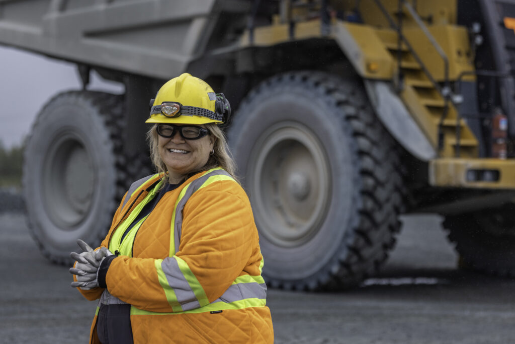 Photo: A woman wearing an orange safety vest and a hard hat is walking to the left of the frame. Behind her you can see part of a large heavy equipment truck used in mining.
