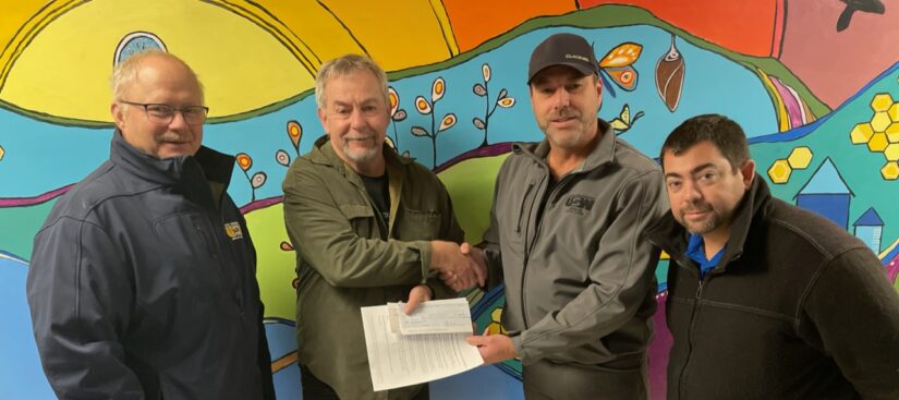 Image: four people stand together in front of a colourful mural backdrop. The two people in the middle are shaking hands as the one second from the right hands a document and a cheque to the person second from left.